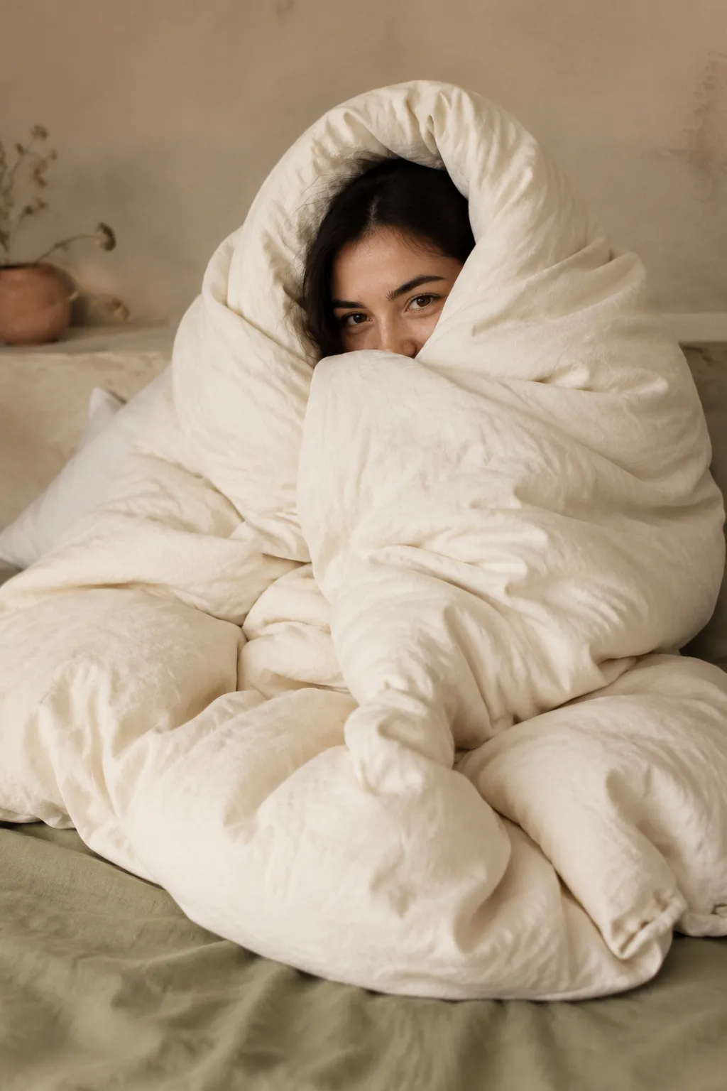 Woman cocooned in a cozy cream comforter against a warm neutral backdrop